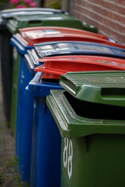 A row of large plastic wheelie bins positioned along a pavement adjacent to a brick wall, with each bin featuring a color-coded lid - green, red, blue, and dark green - indicating different waste type
